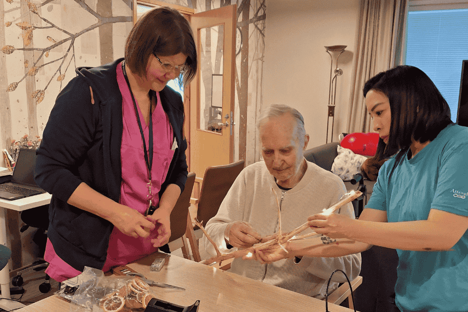 Hoitohenkilökunta avustaa vanhusta käsityöprojektissa hoitokodin yhteisessä tilassa. Pöydällä näkyy tarvikkeita, kuten naruja ja käsityövälineitä