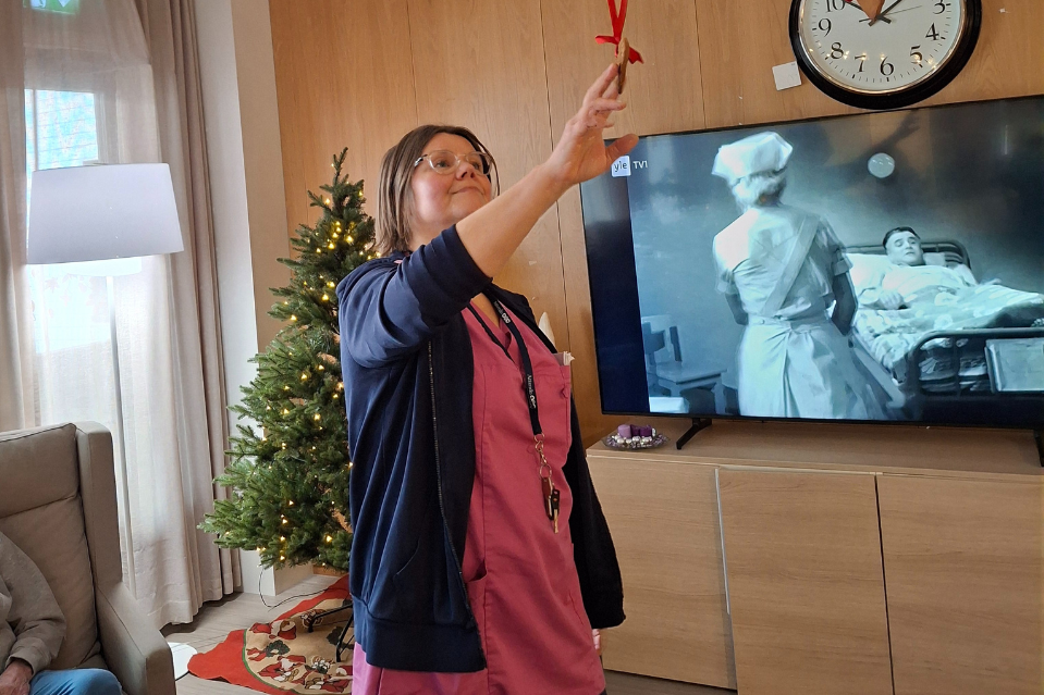 Hoitaja seisoo olohuoneessa joulukuusen vieressä pitäen käsissään punaista koristenauhaa, taustalla televisio.